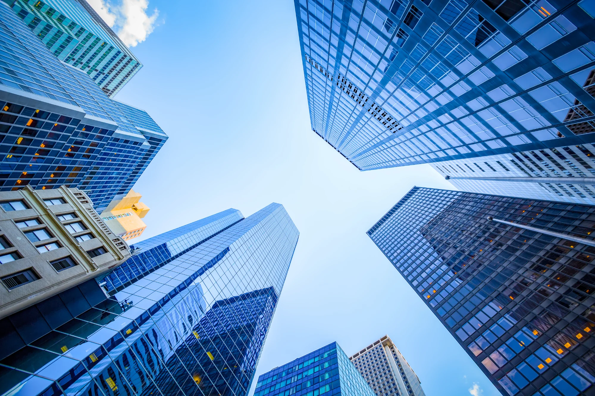 A view looking up from the street level at multiple office towers made of reflective blue glass and white stone, contrasting with the bright blue sky.